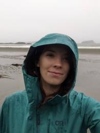 A smiling person stands on a sandy beach in the rain wearing a blue rain jacket with the hood up. She is smiling. 